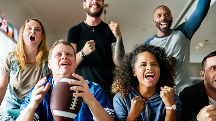 Six male and female football fans cheer their team on at a home watch party.